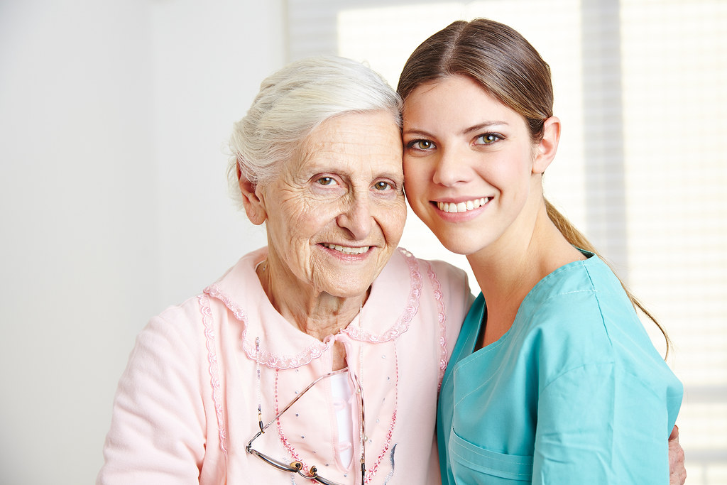 A caregiver in scrubs smiling cheek to cheek with a happy senior woman she cares for